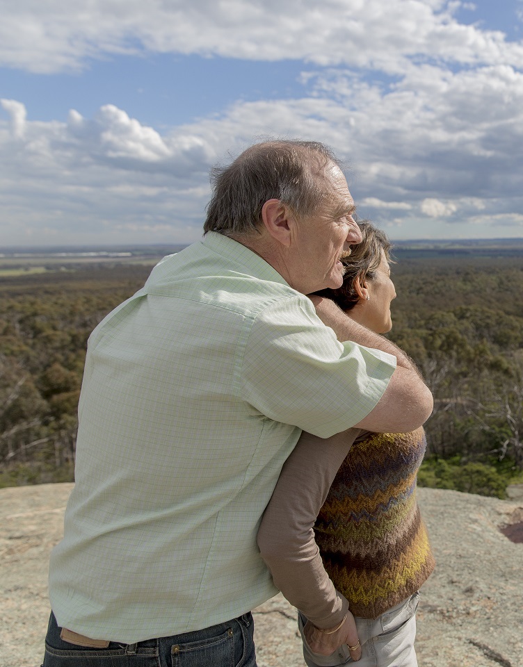 Grey nomad couple embracing on rocky outcrop with bushland landscape background